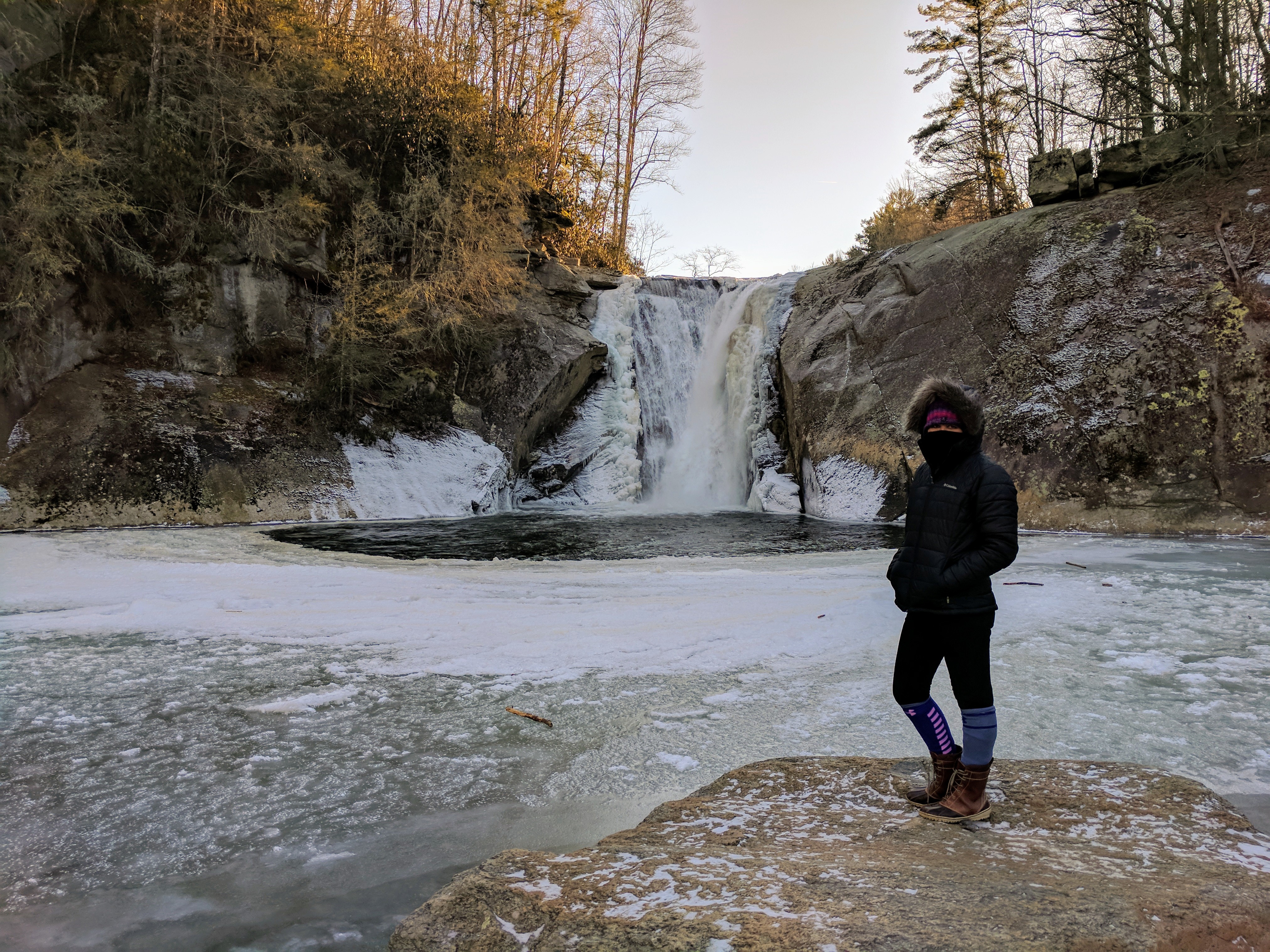 a woman stands on a rock next to a partially frozen pool with icy water pouring in from a nearby waterfall